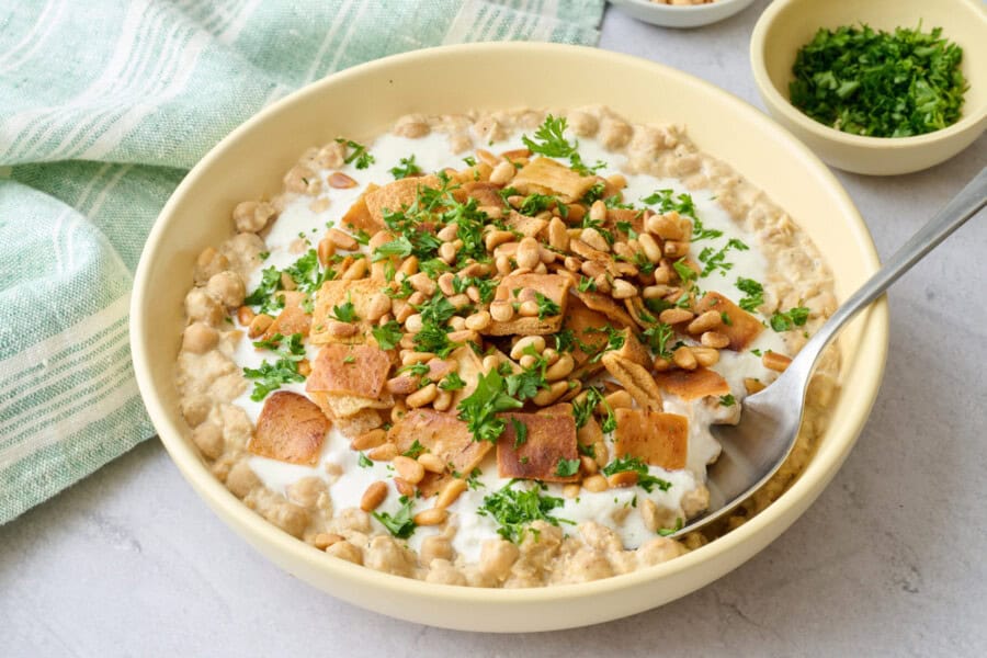 Bowl of chickpea fatteh made with mashed chickpeas, yogurt sauce, toasted pita, and pine nutes.
