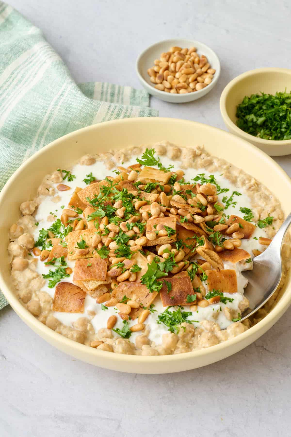 Chickpea fatteh hummus recipe in a bowl layered with mashed chickpeas, yogurt sauce, toasted pita, and pine nuts.