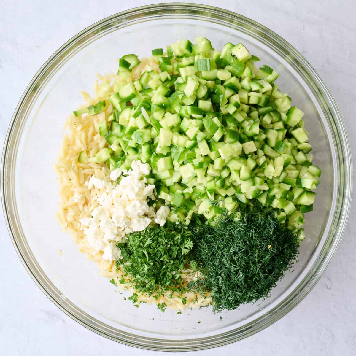 Diced cucumbers, chopped dill and parsley added on top of orzo in a bowl before tossing together.