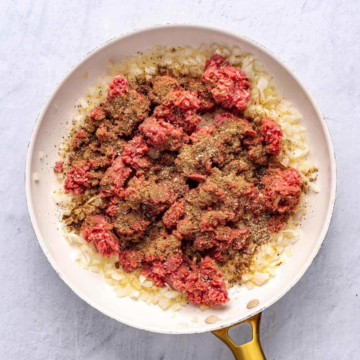 A large skillet of cooked onions and garlic, with ground beef, 7 spice, coriander, salt and pepper added on top.