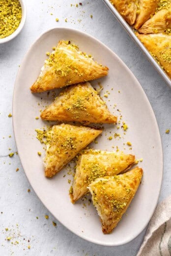 A few Shaabiyat on a long narrow platter garnished with crushed pistachio, baking dish with more still in square shape and a small dish of crushed pistachios nearby.