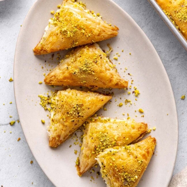 A few Shaabiyat on a long narrow platter garnished with crushed pistachio, baking dish with more still in square shape and a small dish of crushed pistachios nearby.