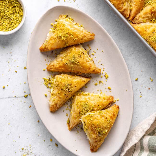 A few Shaabiyat on a long narrow platter garnished with crushed pistachio, baking dish with more still in square shape and a small dish of crushed pistachios nearby.