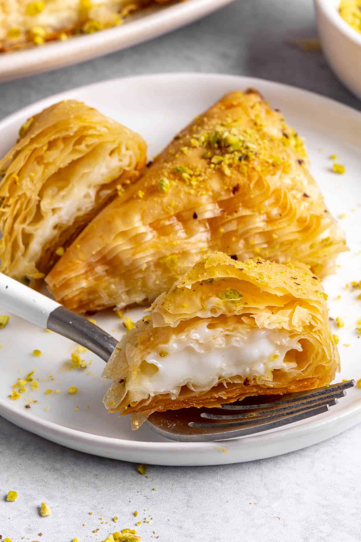2 pieces on a small plate to show flaky phyllo layers and creamy filling, one cut, platter and crushed pistachio dish in background.