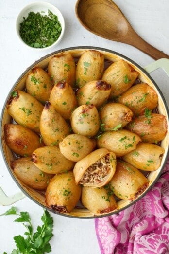 Stuffed onions filled with ground beef, rice and tomatoes in the skillet garnished with chopped parsley, one onion cut open and positioned upwards to see filling inside, small dish of parsley and serving spoon nearby.