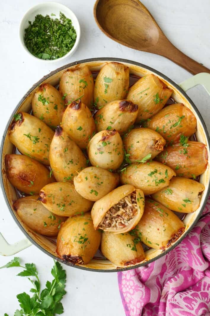 Stuffed onions filled with ground beef, rice and tomatoes in the skillet garnished with chopped parsley, one onion cut open and positioned upwards to see filling inside, small dish of parsley and serving spoon nearby.