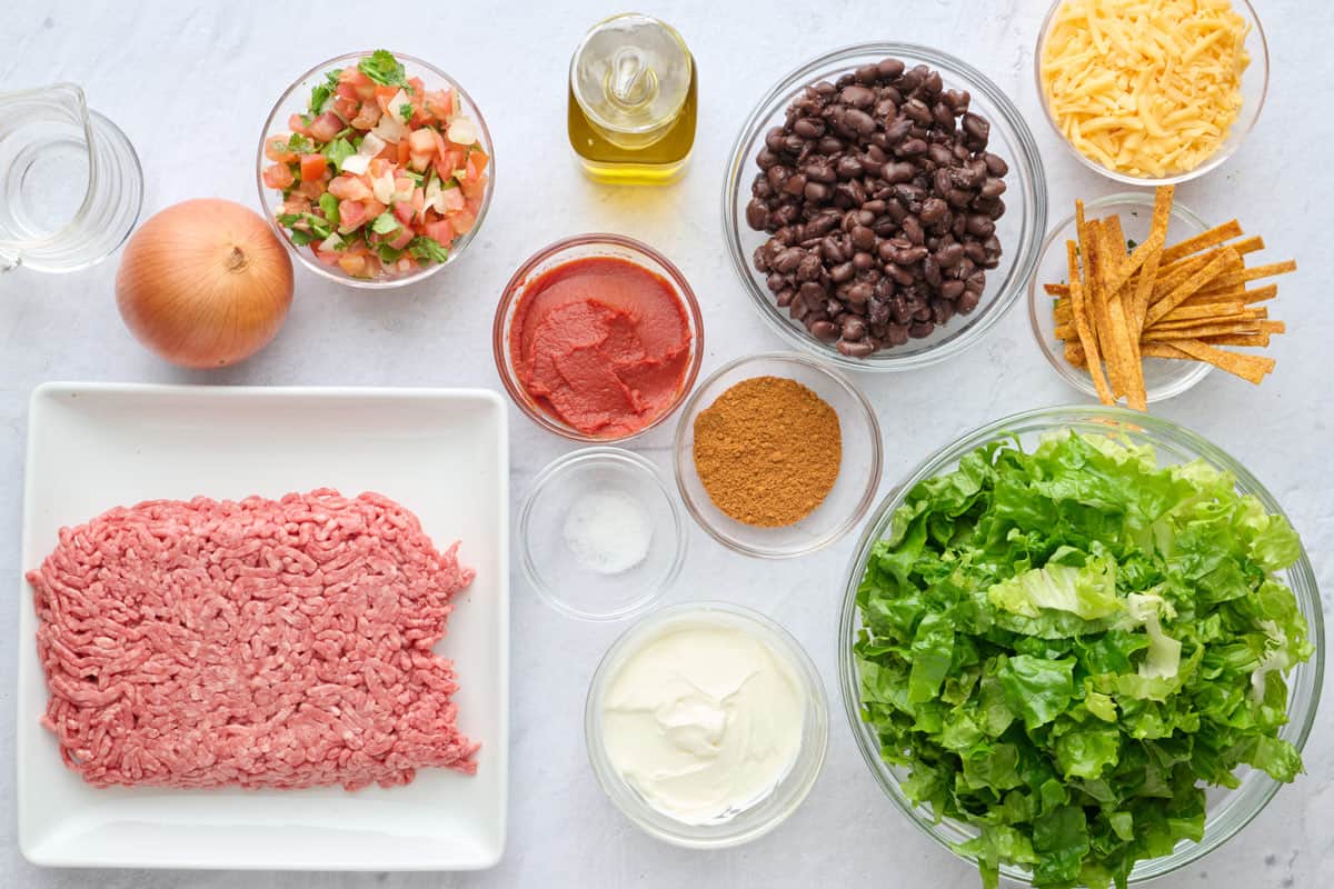 Ingredients for salad recipe: ground beef, onion, pico de gallo, oil, tomato paste, salt, taco seasoning, sour cream, black beans, tortilla strips, shredded cheese, and chopped lettuce.