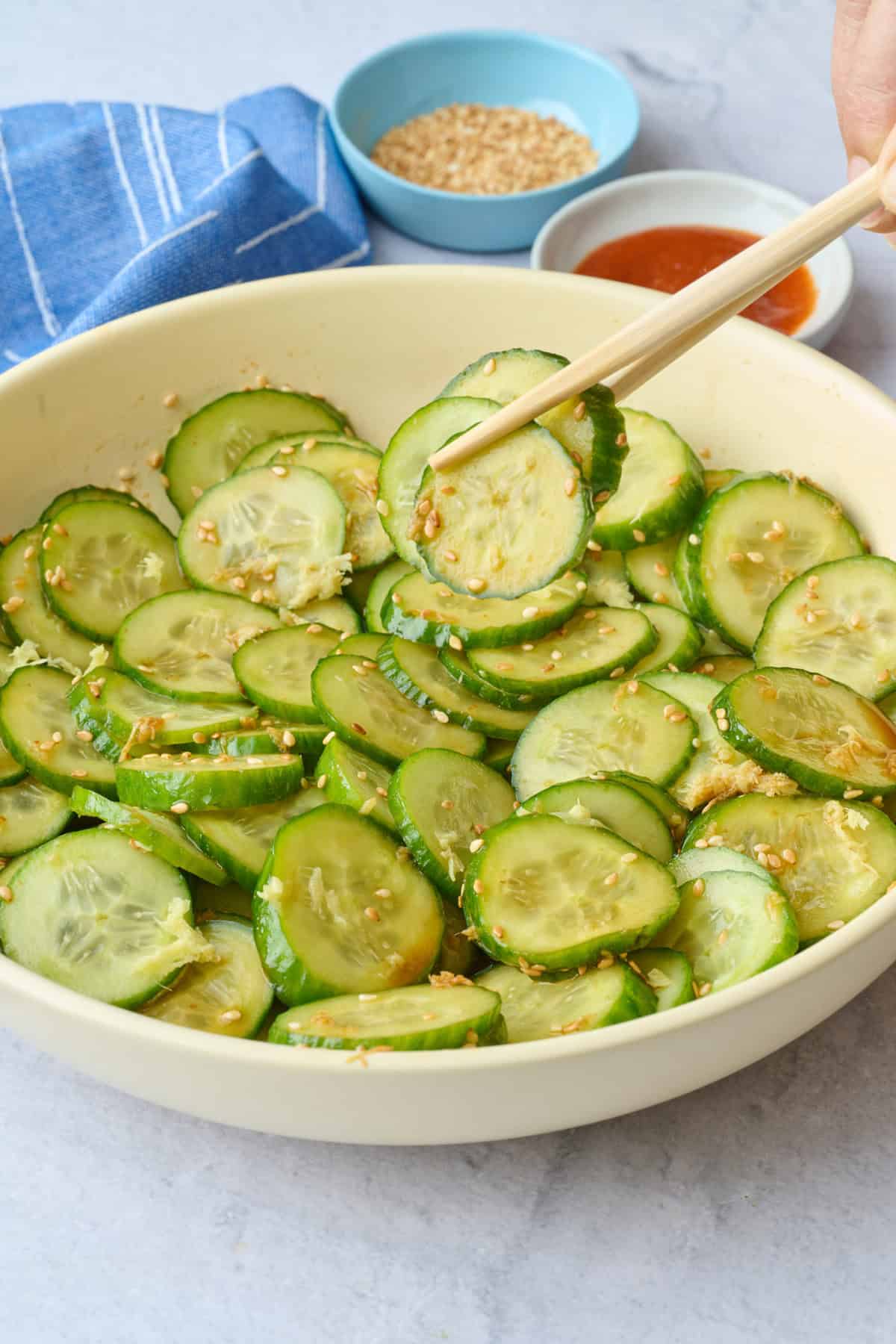 Recette de salade de concombre à l'asiatique 12 Salade de concombre asiatique dans un bol de service avec des graines de sésame saupoudrées sur le dessus, ainsi que de petites assiettes de graines de sésame supplémentaires et des baguettes sur le dessus du bol.