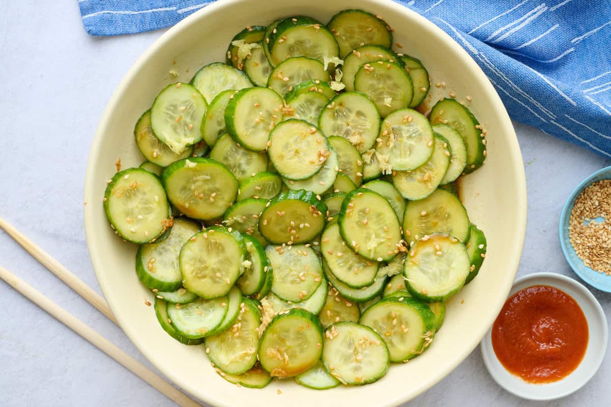 Recette de salade de concombre à l'asiatique 3 Salade de concombre asiatique dans un bol de service avec des graines de sésame saupoudrées sur le dessus, et de petites assiettes de graines de sésame supplémentaires et des baguettes à proximité.