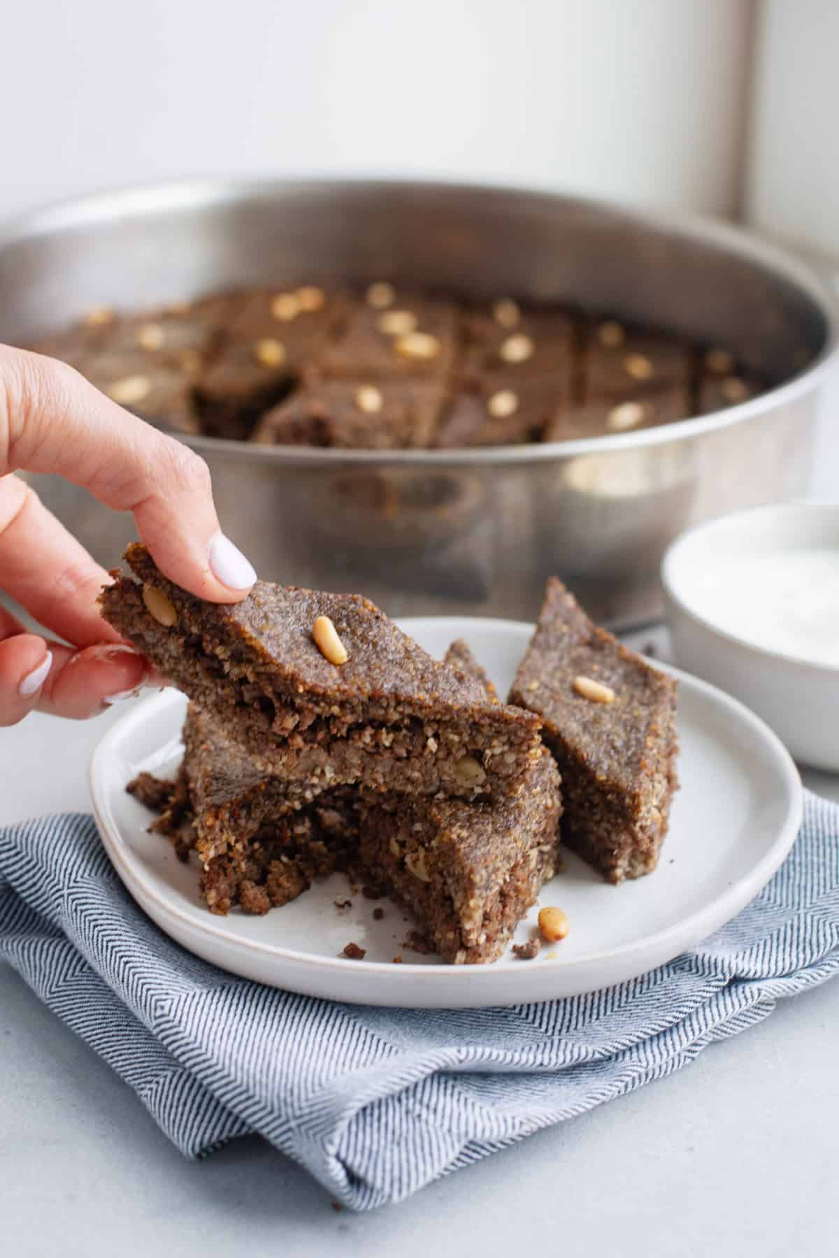 Hand lifting up a diamond sliced piece of baked beef kibbeh from a plate with more pieces.