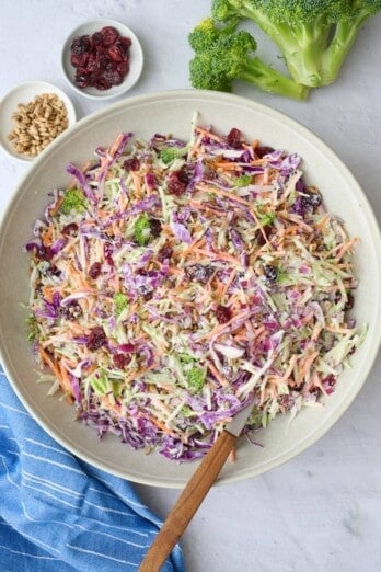 Broccoli slaw in a serving bowl with serving set dipped inside, small dishes of extra dried cranberries and sunflower seeds nearby.