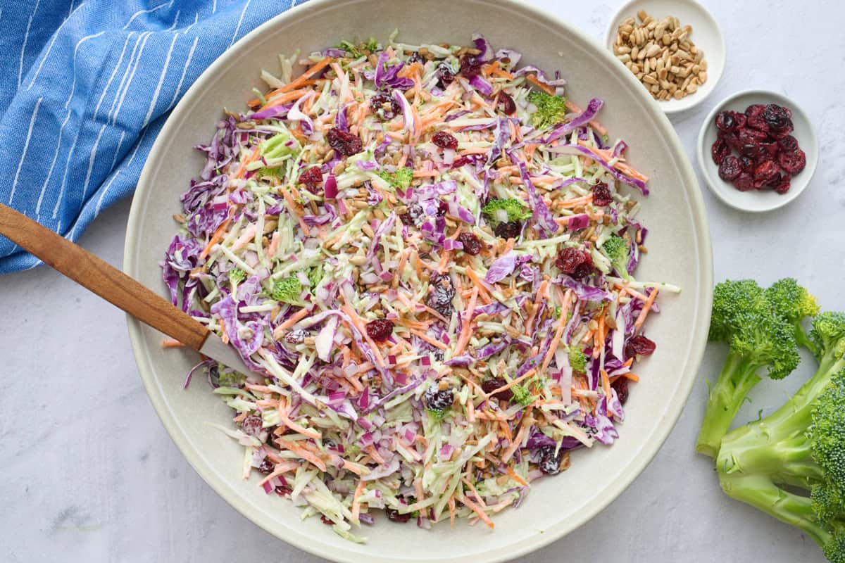 Broccoli slaw in a serving bowl with serving set dipped inside, small dishes of extra dried cranberries and sunflower seeds nearby.