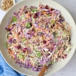 Broccoli slaw in a serving bowl with serving set dipped inside, small dishes of extra dried cranberries and sunflower seeds nearby.