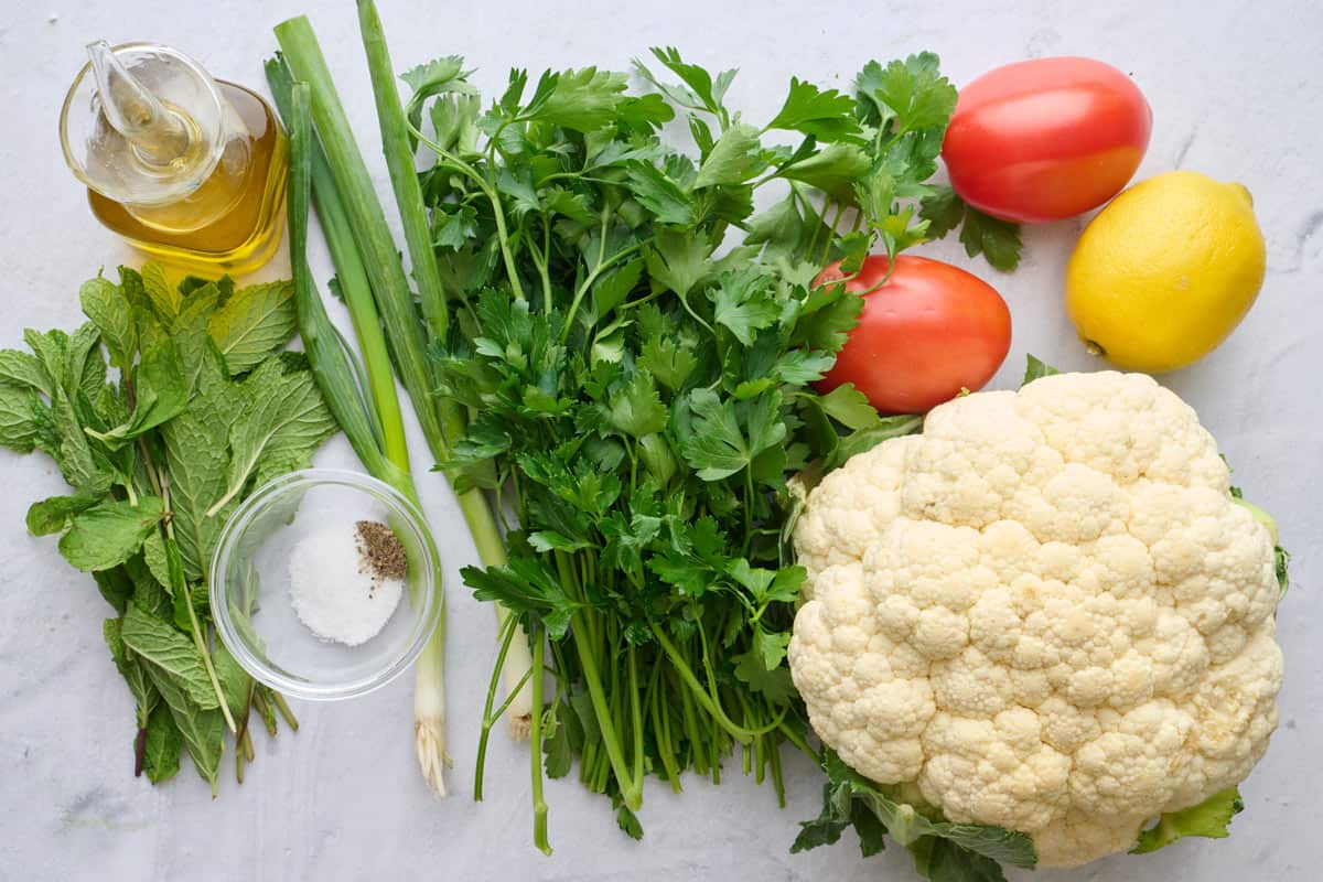 Ingredients for tabbouleh salad recipe made without bulgur: cauliflower, parsley, green onions, mint, lemon tomatoes, oil, salt and pepper.