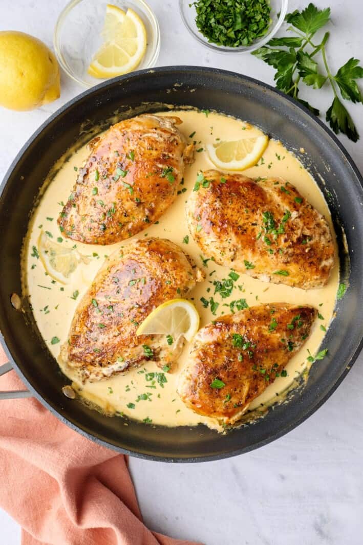 Creamy lemon chicken in skillet with sauce spooned lightly on top of each chicken, garnished with chopped parsley, small dish of lemons nearby.