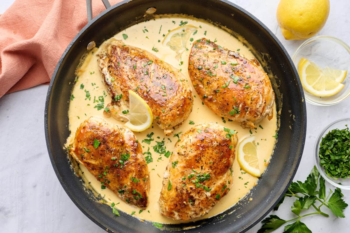 Creamy lemon chicken in skillet with sauce spooned lightly on top of each chicken, garnished with chopped parsley, small dish of lemons nearby.