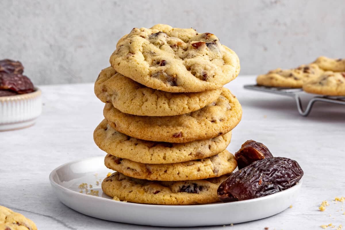 Soft and chewy date cookies stacked on a plate with whole dates and wire rack of more cookies nearby.