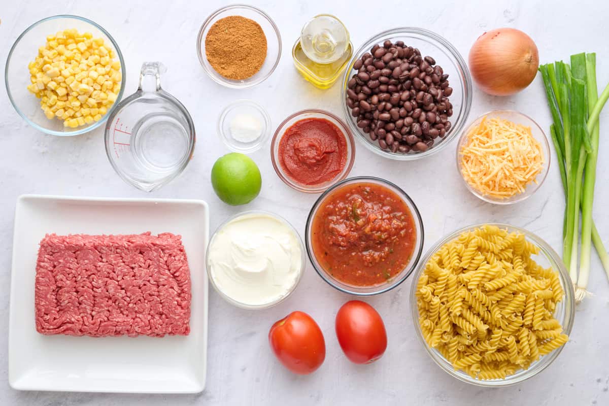 Ingredients for taco pasta salad recipe: ground beef, corn, taco seasoning, salt, lime, sour cream, tomato paste, oil, black beans, shredded cheese, onion, green onions, tomatoes, salsa, and dry pasta.