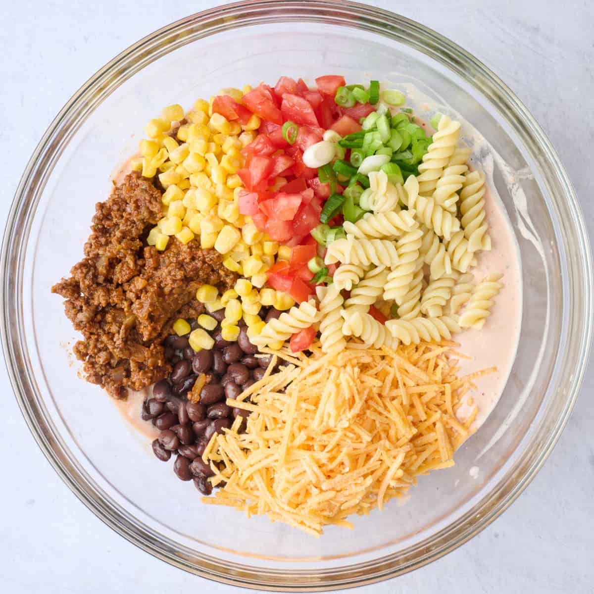 Cooked pasta, taco meat, corn, tomatoes, green onions, beans, and shredded cheese added on top of creamy sauce before tossing together.