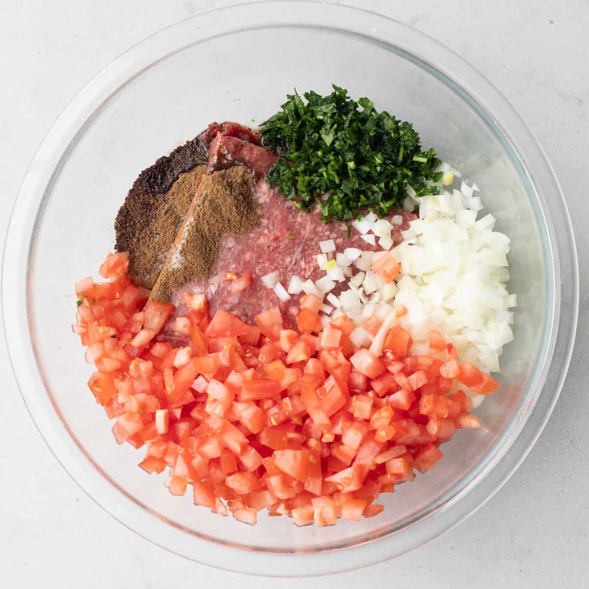 Ground beef, spices, chopped parsley, onions, and tomatoes in a bowl before combining.