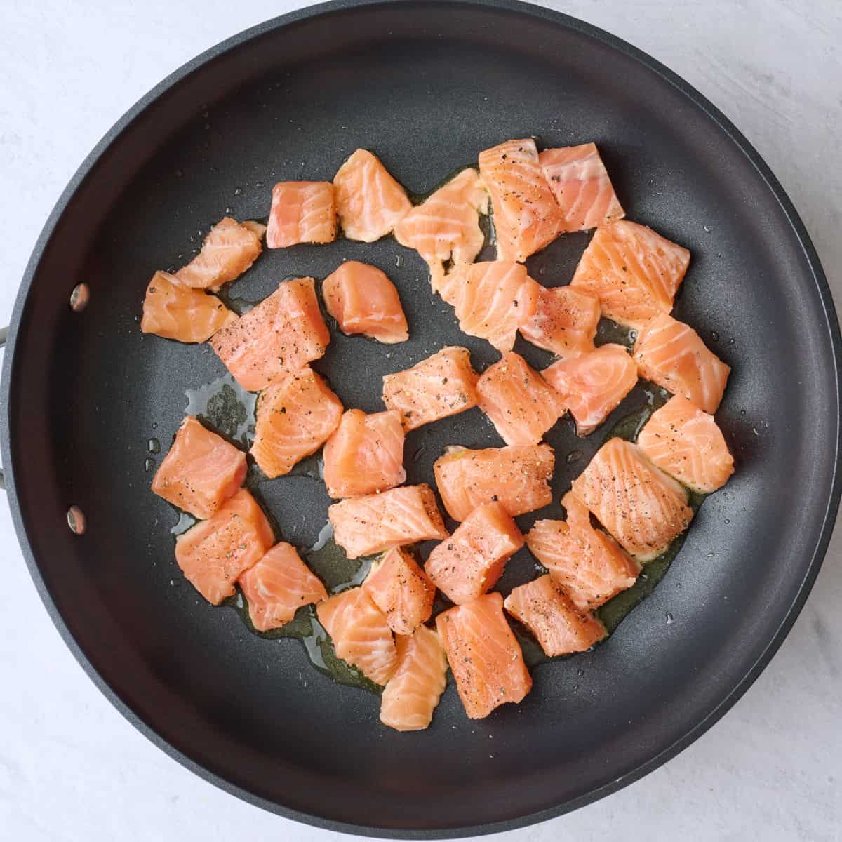 Seasoned salmon cubes in a large oiled skillet before cooking.
