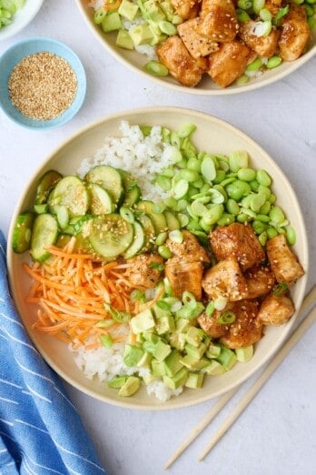 Two salmon bowls assembled with cucumber salad, carrots, edamame, and avocado garnished with a light sprinkle of sesame seeds, green onions, and a drizzle of sriracha.
