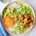 Two salmon bowls assembled with cucumber salad, carrots, edamame, and avocado garnished with a light sprinkle of sesame seeds, green onions, and a drizzle of sriracha.