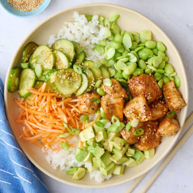 Two salmon bowls assembled with cucumber salad, carrots, edamame, and avocado garnished with a light sprinkle of sesame seeds, green onions, and a drizzle of sriracha.