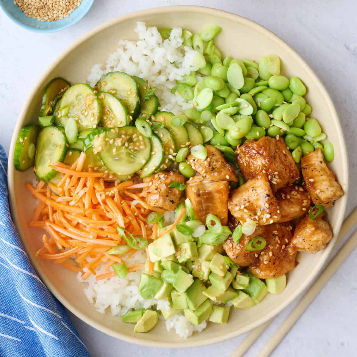Two salmon bowls assembled with cucumber salad, carrots, edamame, and avocado garnished with a light sprinkle of sesame seeds, green onions, and a drizzle of sriracha.