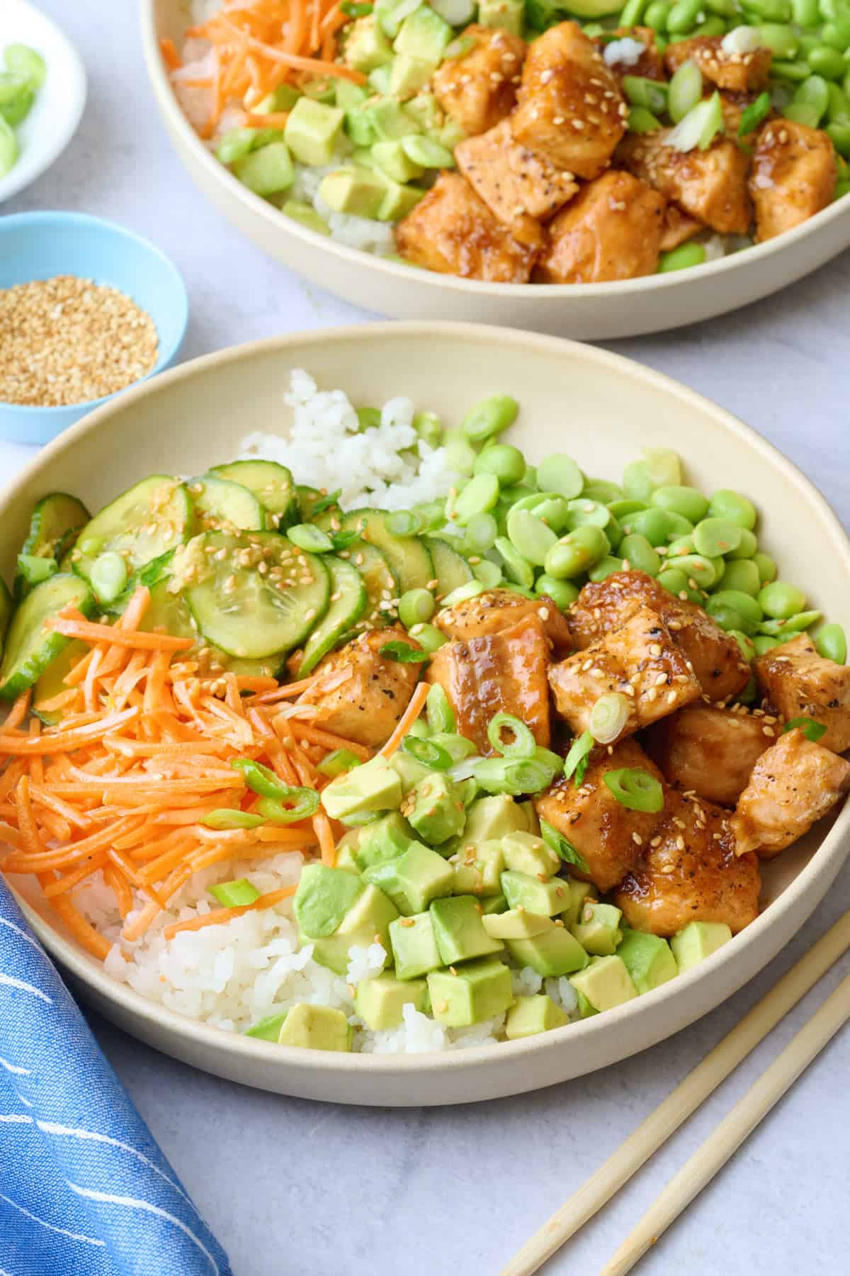Two salmon bowls assembled with cucumber salad, carrots, edamame, and avocado garnished with a light sprinkle of sesame seeds, green onions, and a drizzle of sriracha.