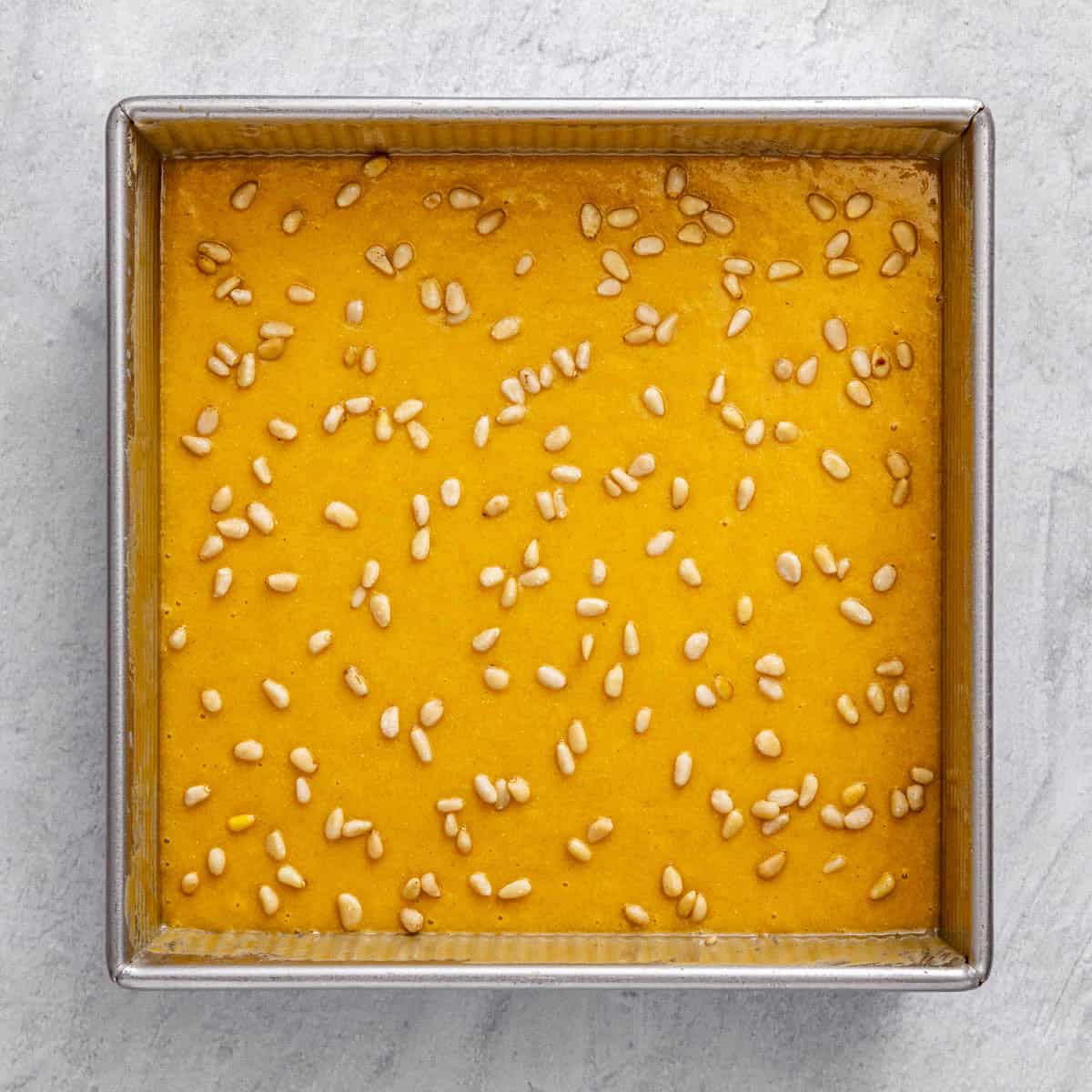 Cake batter in the tahini brushed square baking pan with pine nuts on top before baking.