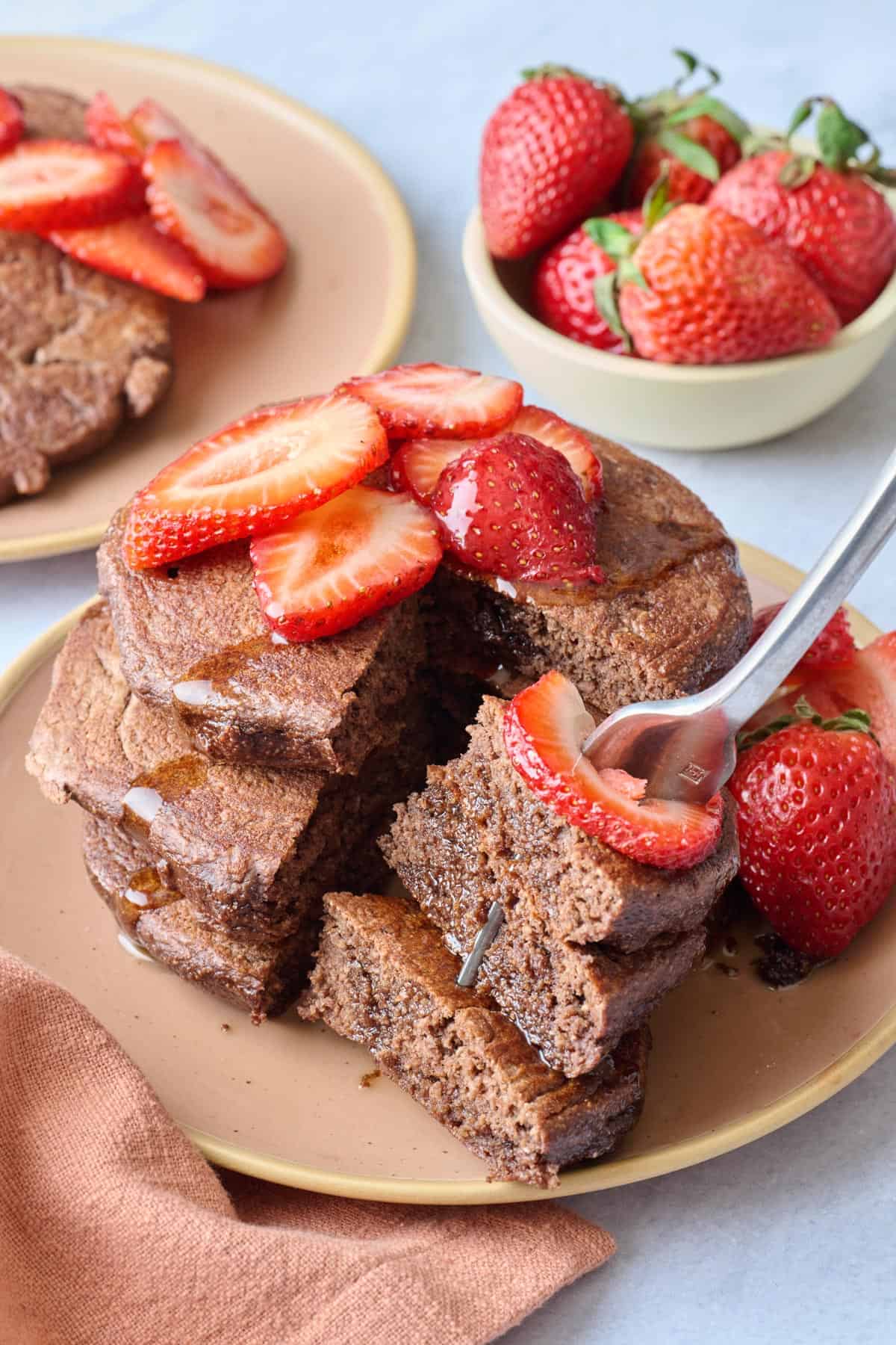 Fork lifting up a bite of the best chocolate protein pancakes from stack with strawberries and maple syrup.