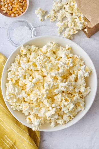 Popcorn in a bowl with the paper bag, laying down with some pouring out onto the surface, small dish of salt nearby.