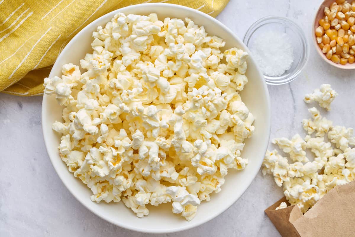 Popcorn in a bowl with the paper bag, laying down with some pouring out onto the surface, small dish of salt nearby.