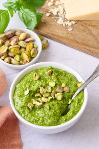 Pistachio pesto in a small bowl with a spoon dipped inside, garnished with a few chopped pistachios, extra fresh ingredients around.