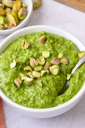 Pistachio pesto in a small bowl with a spoon dipped inside, garnished with a few chopped pistachios, extra fresh ingredients around.