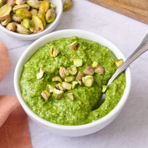 Pistachio pesto in a small bowl with a spoon dipped inside, garnished with a few chopped pistachios, extra fresh ingredients around.