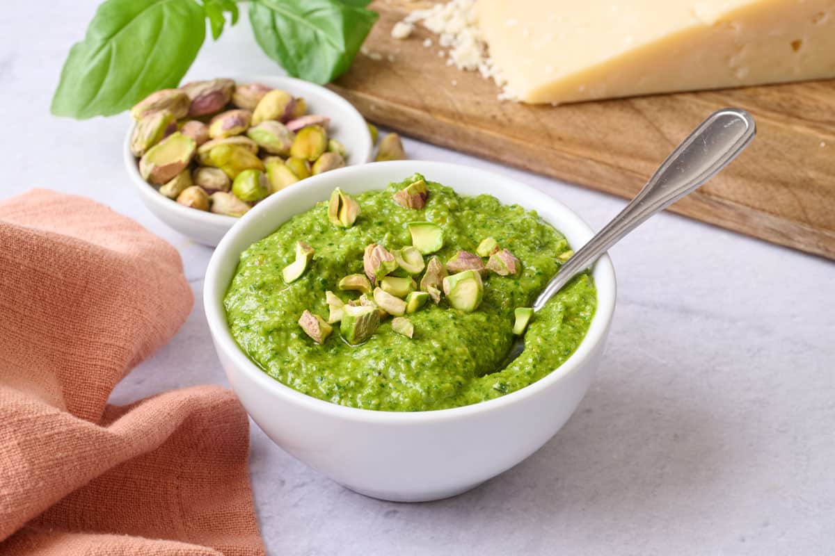 Pistachio pesto in a small bowl with a spoon dipped inside, garnished with a few chopped pistachios, extra fresh ingredients around.