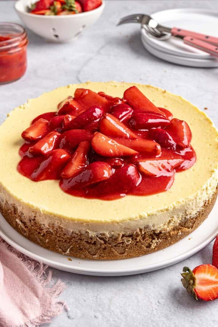 Whole strawberry cheesecake on a round platter with a small dish of extra strawberry sauce nearby.