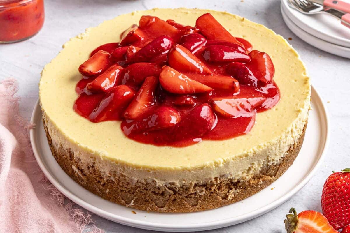 Whole strawberry cheesecake on a round platter with a small dish of extra strawberry sauce nearby.