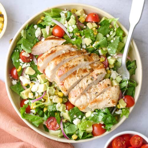 2 bowls of grilled chicken salad with extra fresh ingredients around.
