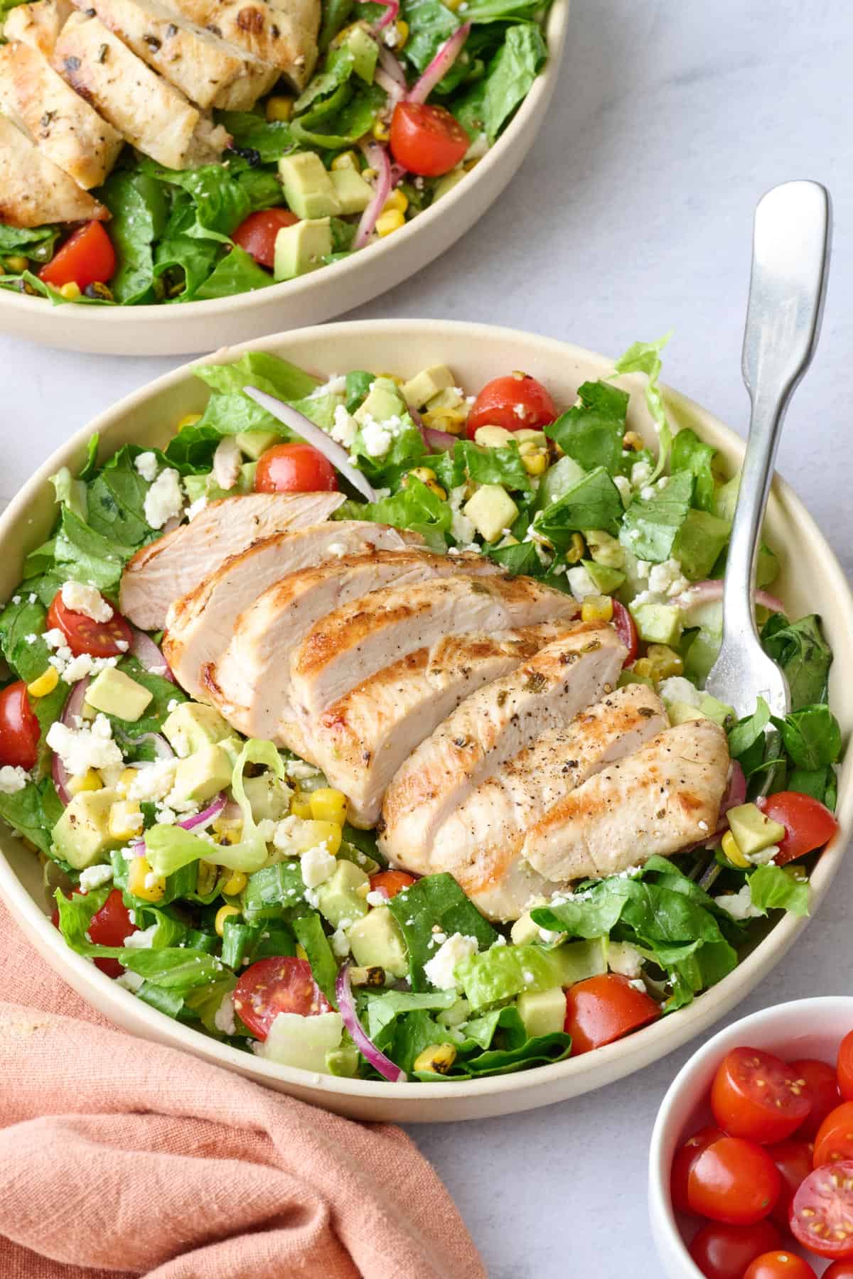2 bowls of grilled chicken salad with extra fresh ingredients around.
