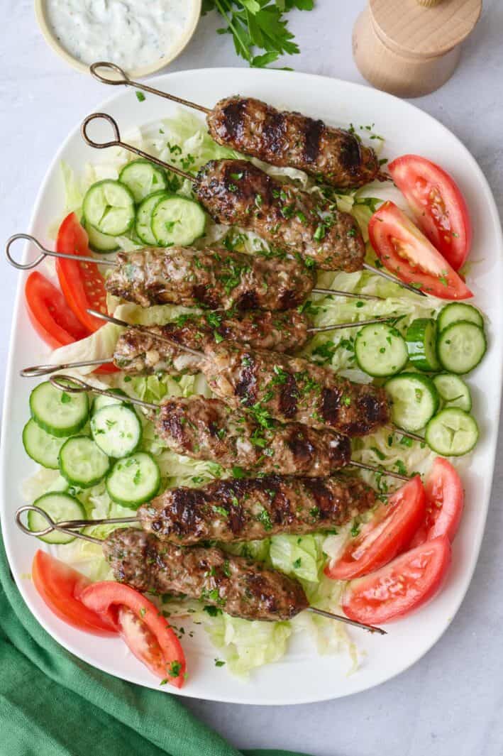 Ground lamb kabobs on skewers on a large narrow platter on top of shredded lettuce with wedges of tomatoes and slices of cucumber scattered yogurt sauce nearby, garnished with chopped parsley.