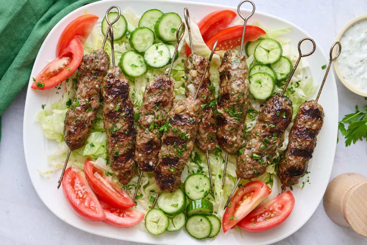 Ground lamb kabobs on skewers on a large narrow platter on top of shredded lettuce with wedges of tomatoes and slices of cucumber scattered yogurt sauce nearby, garnished with chopped parsley.