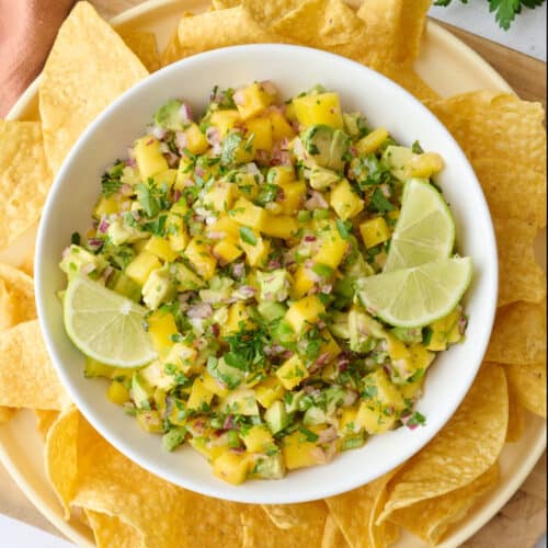 Mango salsa in a serving bowl garnished with lime wedges and extra cilantro, tortilla chips and a halved mango cut on the skin.