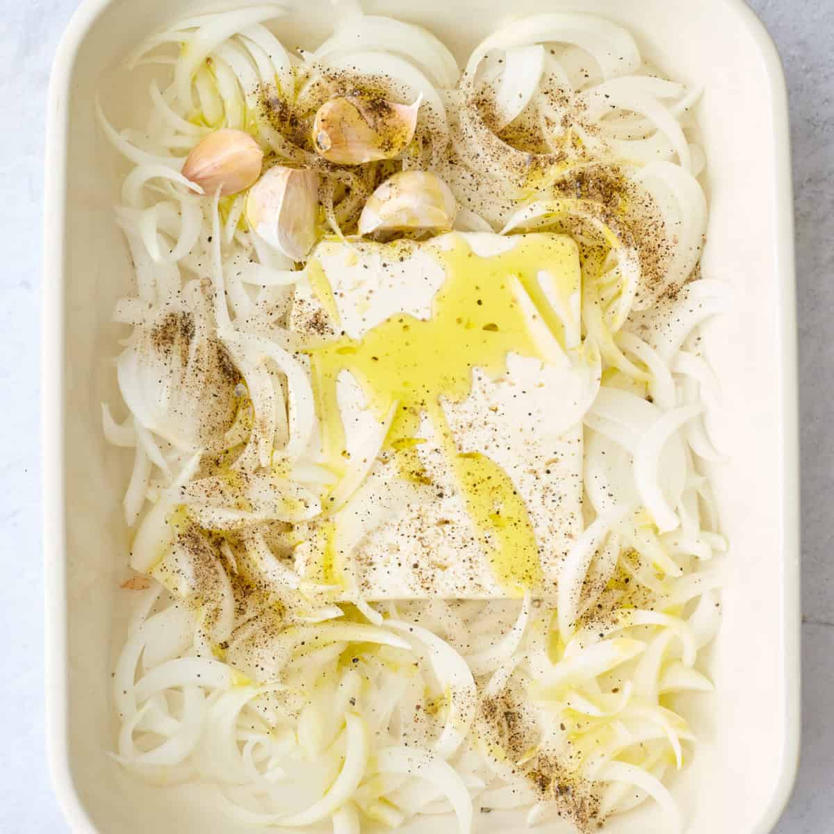 Feta block in a baking dish with sliced onions and garlic around, drizzled with olive oil and seasoned with pepper before baking.