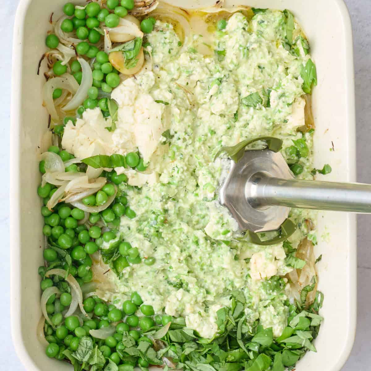 Peas, cooked onions, feta, and garlic in a baking dish being blended with an immersion blender.