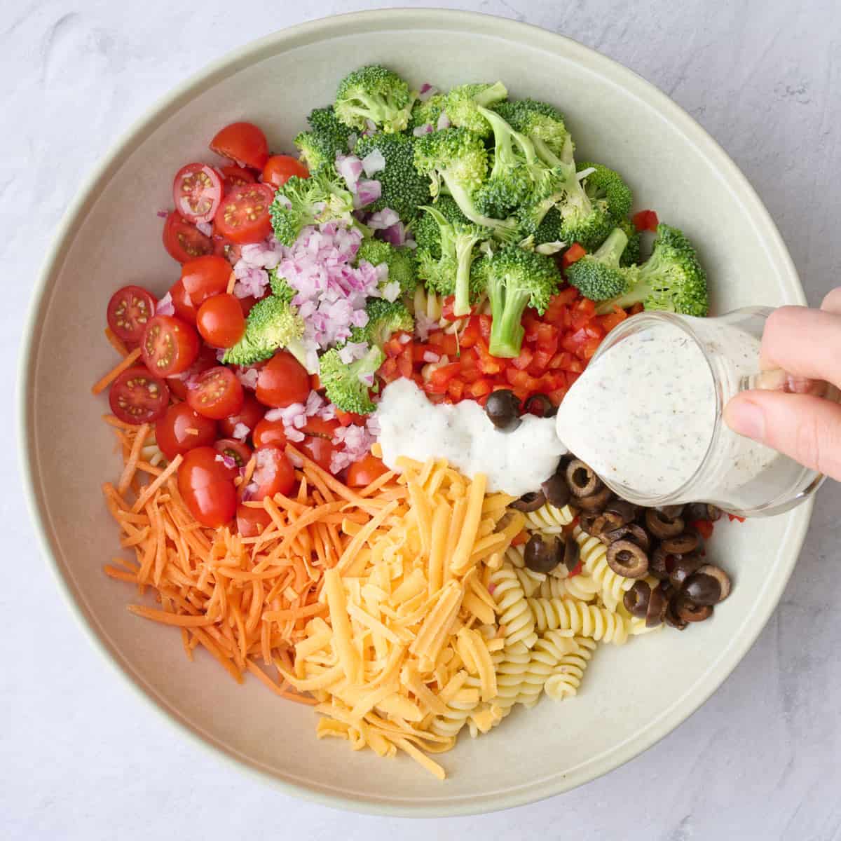 Cooled pasta, tomatoes, broccoli, pepper, carrots, cheese, onions, and olive in a large bowl before tossing with dressing being poured on top.