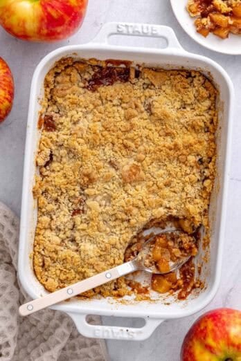 Apple crumble in the baking dish with serving removed to a small plate nearby, serving spoon dipped inside, a few apples around.