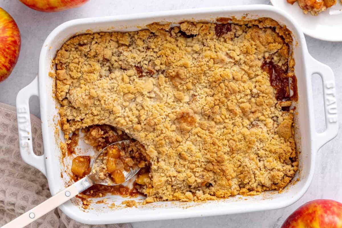 Apple crumble in the baking dish with serving removed to a small plate nearby, serving spoon dipped inside, a few apples around.